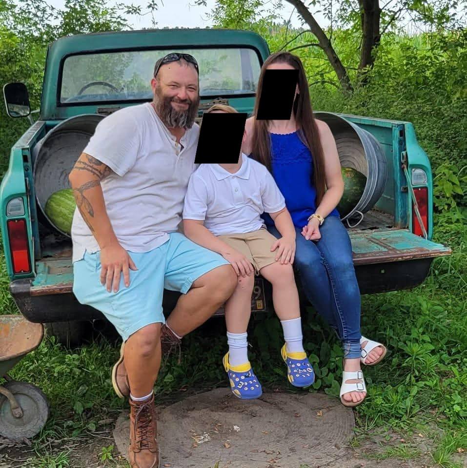 Family portrait featuring a smiling man, woman, and child sitting on the back of a vintage green truck surrounded by greenery.