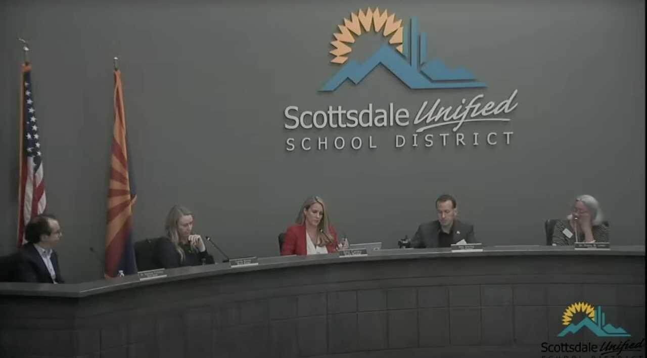 Scottsdale Unified School District board members discuss educational policies during a meeting, with flags displayed in the background.