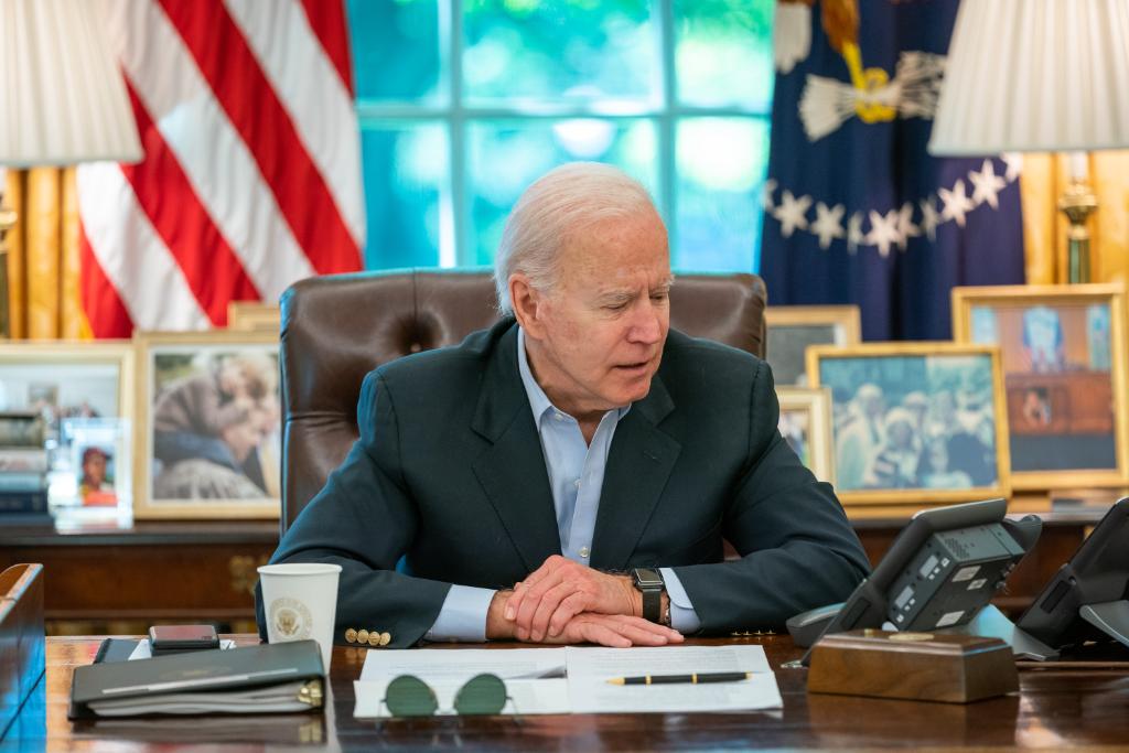 President Joe Biden sitting at the Resolute Desk in the Oval Office, engaged in a phone conversation with the American flag in the background.
