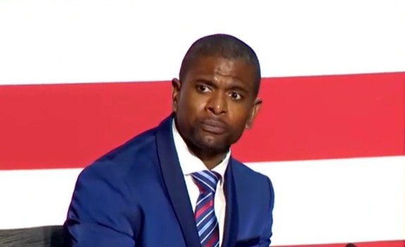 Man in a blue suit with a concerned expression sits against a backdrop of red and white stripes, conveying a serious moment during a discussion or event.