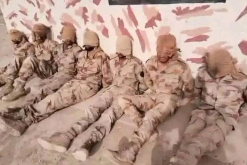 Soldiers in camouflage uniforms sitting against a wall, with their heads covered, in a sandy environment.