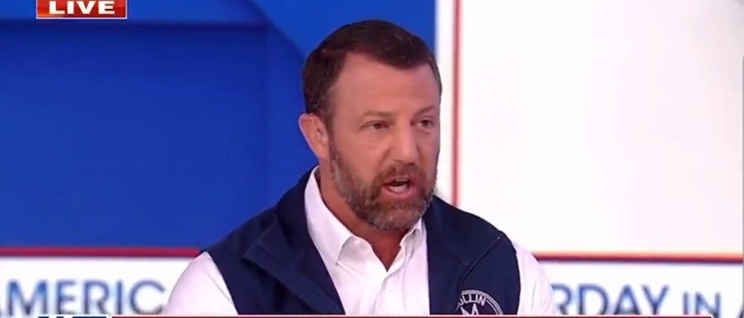 Man speaking passionately during a live television segment, wearing a blue vest over a white shirt, with a colorful background.