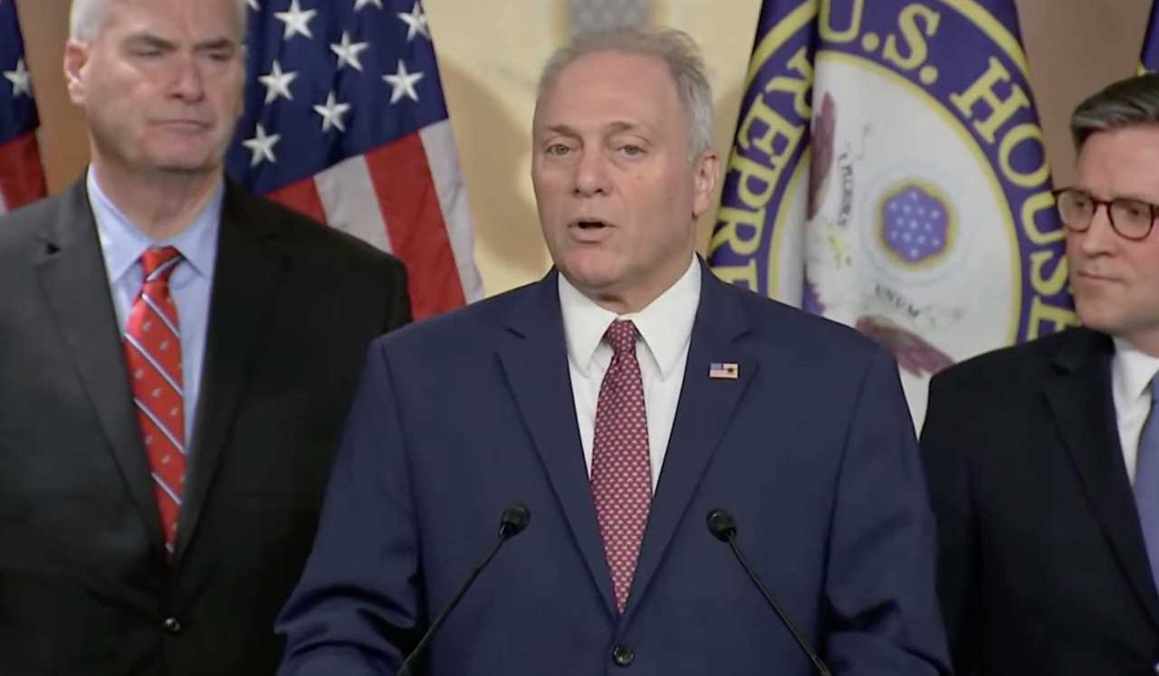 U.S. House Speaker addresses the media during a press conference, flanked by colleagues and American flags in the background.