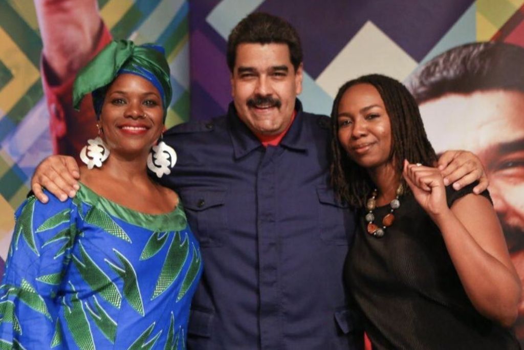 Nicolas Maduro poses with two women, one in traditional attire, at a vibrant event backdrop featuring geometric patterns.