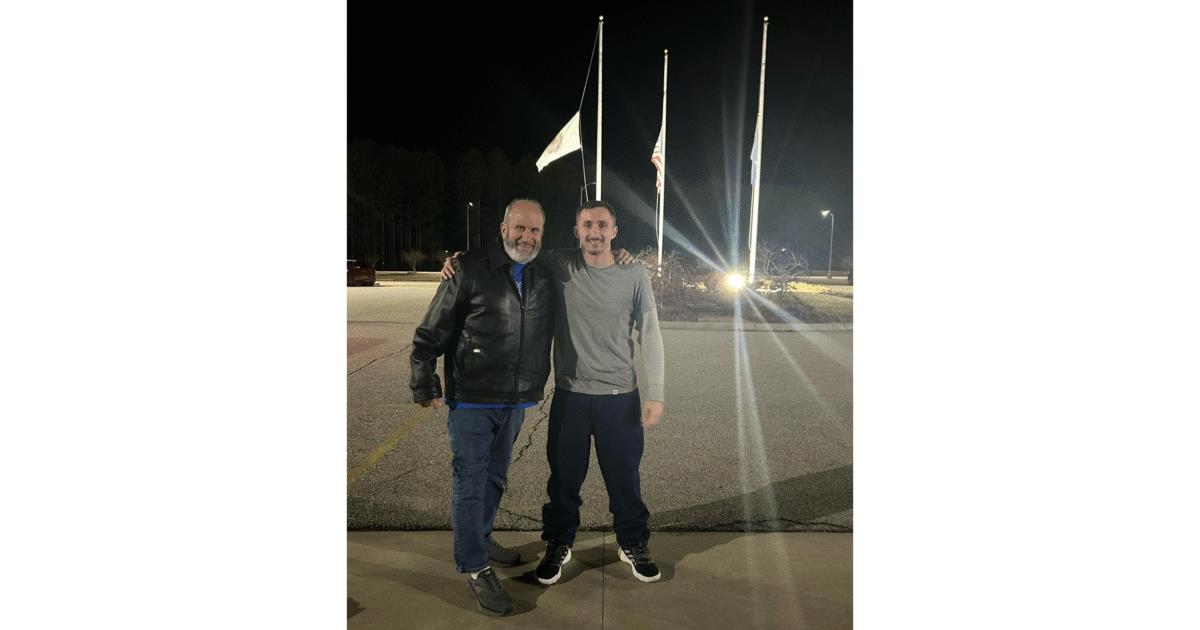 Two men pose together in a parking lot at night, with flags at half-mast in the background, creating a warm and friendly atmosphere.