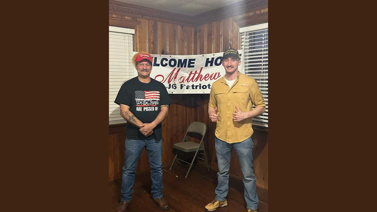 Two men pose together in a warmly decorated room, celebrating a welcome home event with a banner for Matthew, a J6 patriot.