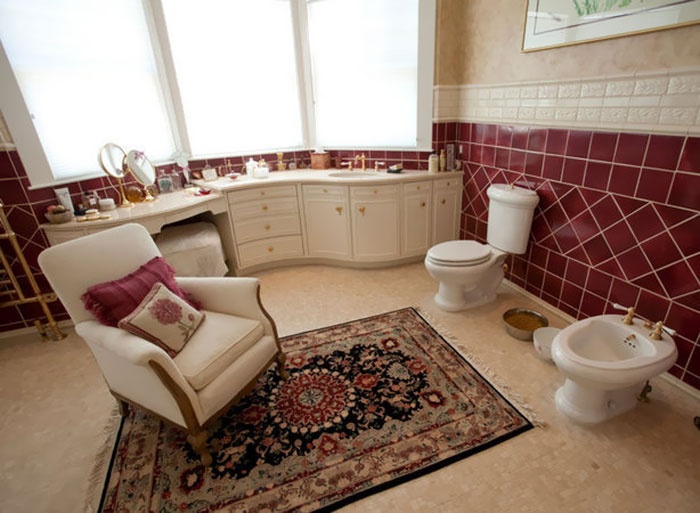 Ivory and burgundy primary bathroom with two toilets, a rug, chair, vanity, sink and windows.