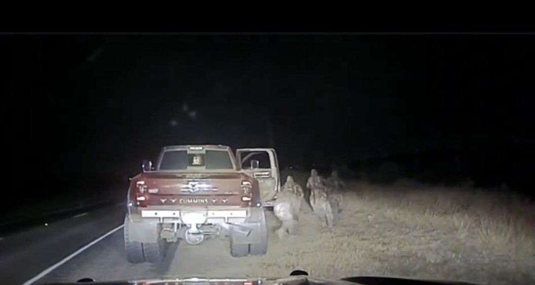 Nighttime scene showing a red truck parked on the side of a road with individuals in camouflage approaching the vehicle.