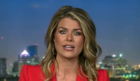 News anchor with long blonde hair wearing a red blazer, presenting against a city skyline backdrop at dusk.