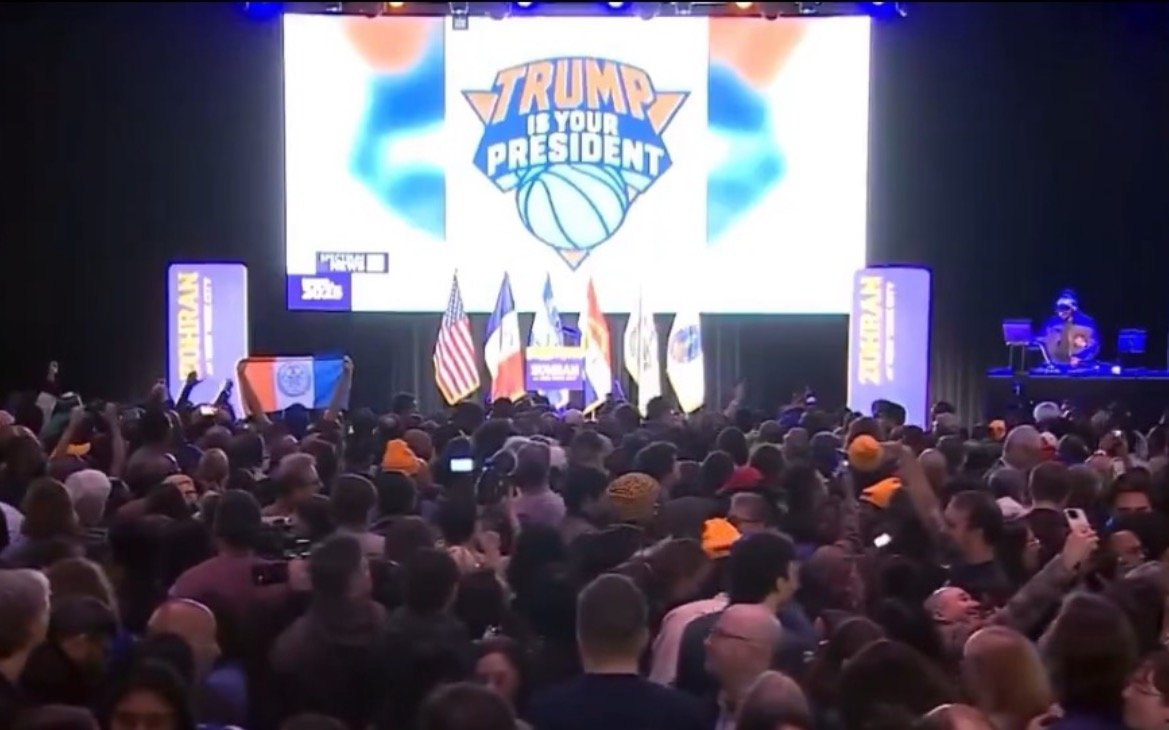 Crowd gathered at a political event featuring a large "Trump is Your President" banner, with flags and a DJ in the background.