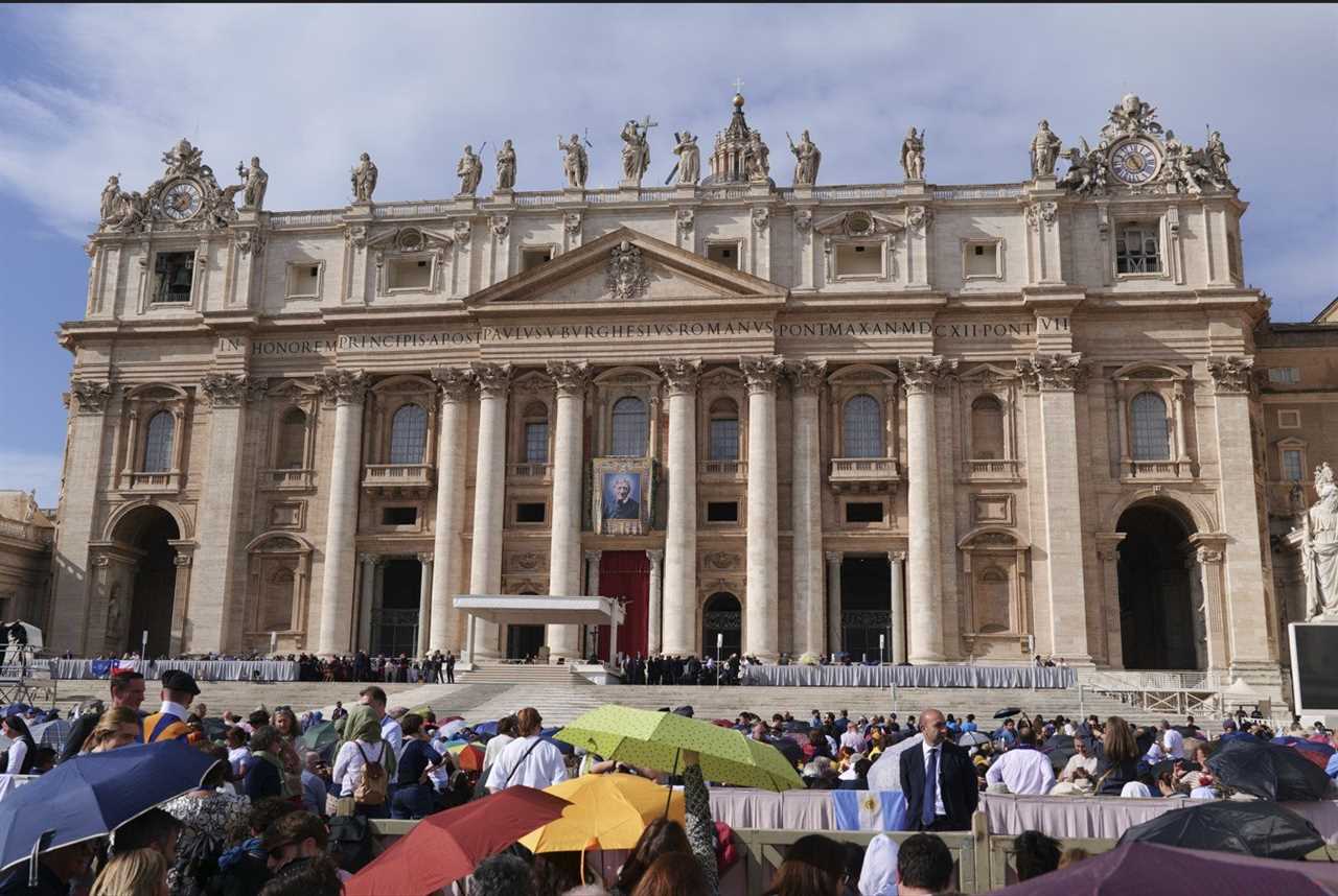 St. Peter's Basilica in Vatican City, showcasing its grand façade and a large gathering of visitors with umbrellas on a sunny day.