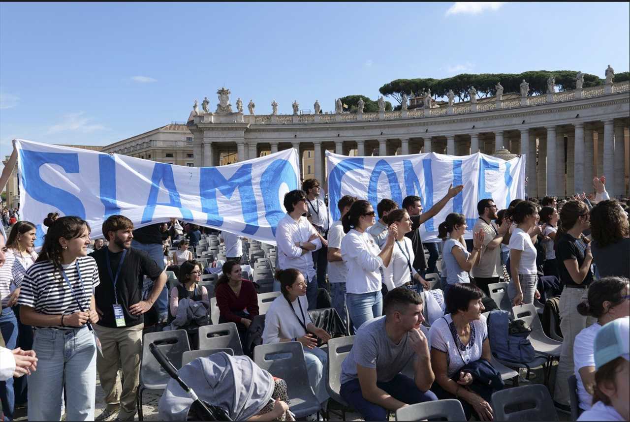 Crowd holding a large banner reading "SIAMO CON LEI" at a public event in a historic plaza, with seated audience in the background.