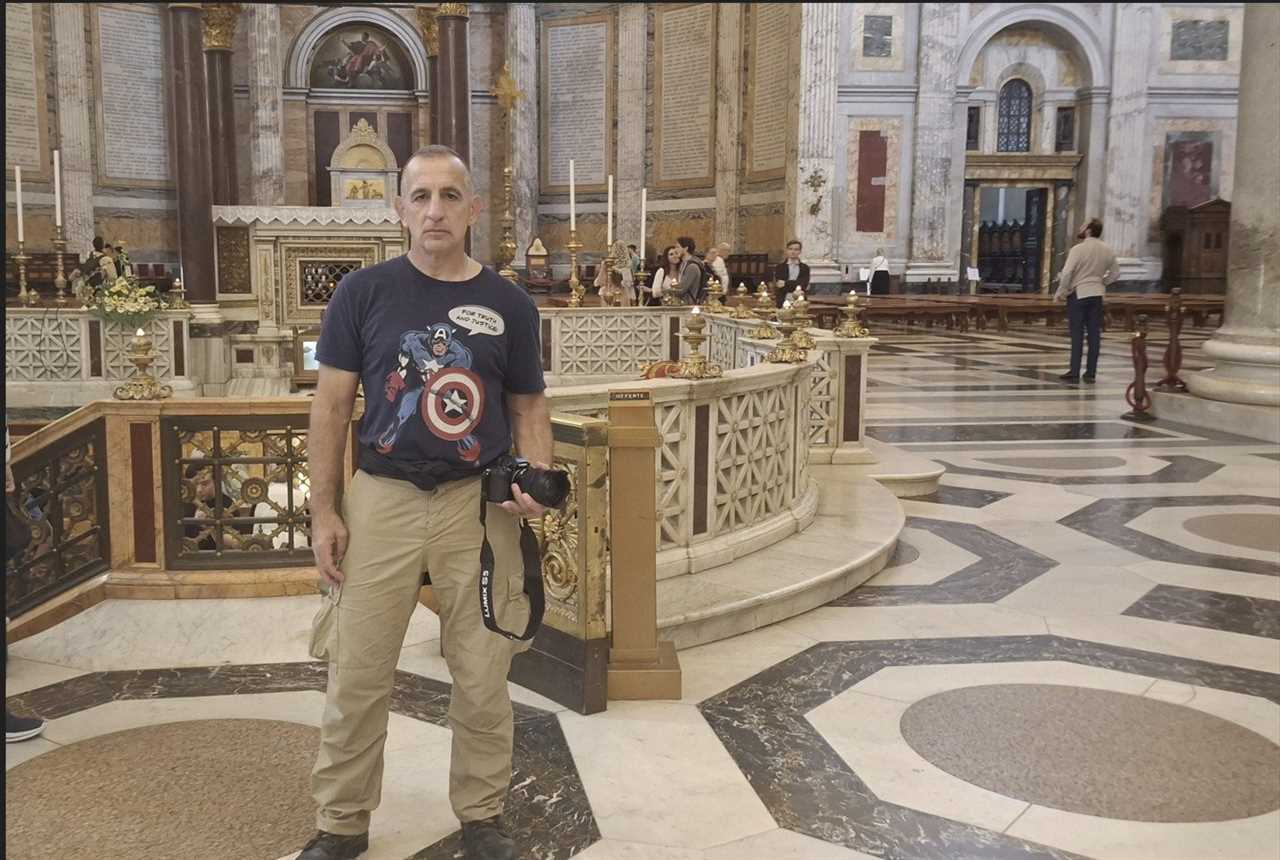 Man in a Captain America t-shirt holding a camera stands in a grand, ornate church interior with marble floors and historical architecture.