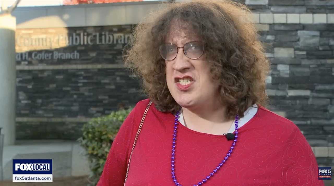 Person speaking outside a public library, wearing a red shirt and glasses, expressing strong emotions during an interview.