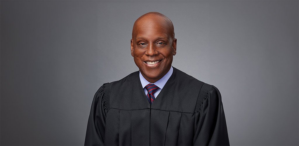 Portrait of a smiling male judge wearing a black robe and a striped tie against a neutral background.