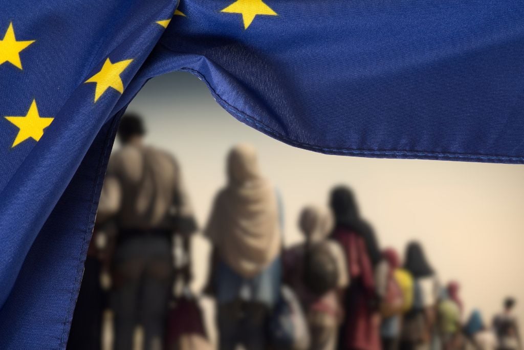European Union flag in the foreground with a blurred line of migrants in the background, symbolizing migration and refugee issues in Europe.