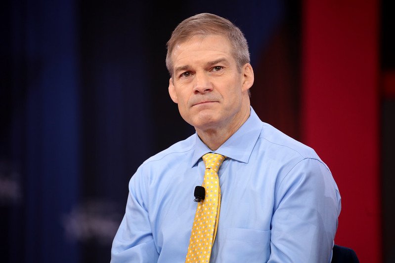 Man in a blue shirt and yellow tie sitting on stage, looking thoughtfully during a discussion at a conference.