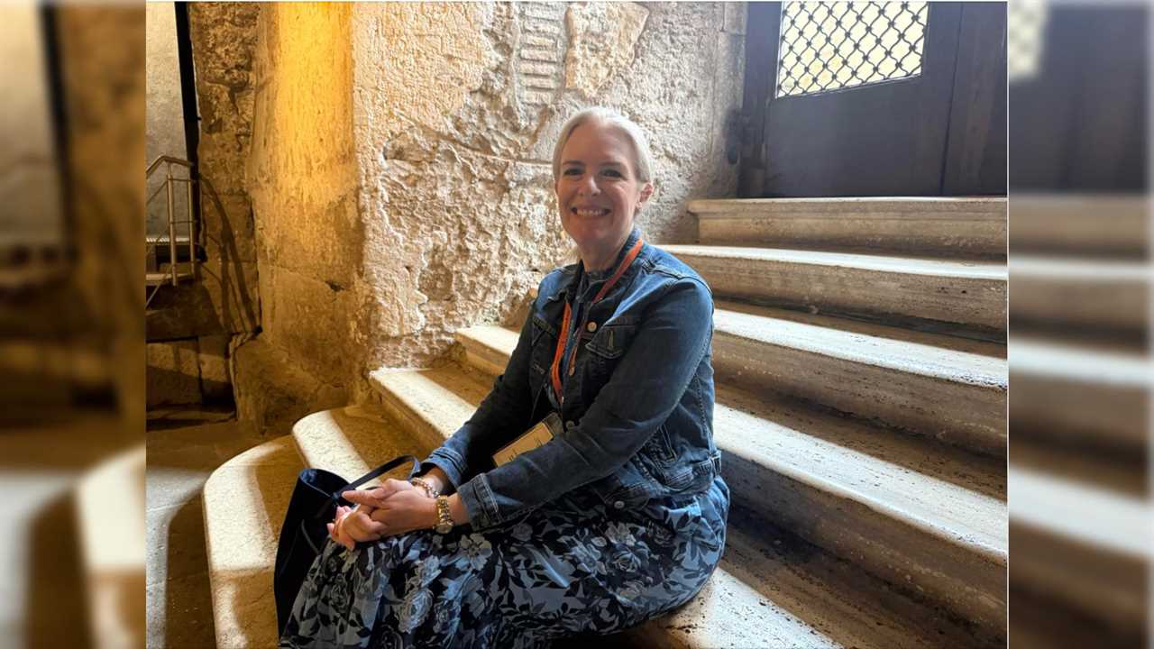 Smiling woman in a denim jacket sitting on stone steps, showcasing a historic interior with warm lighting and textured walls.