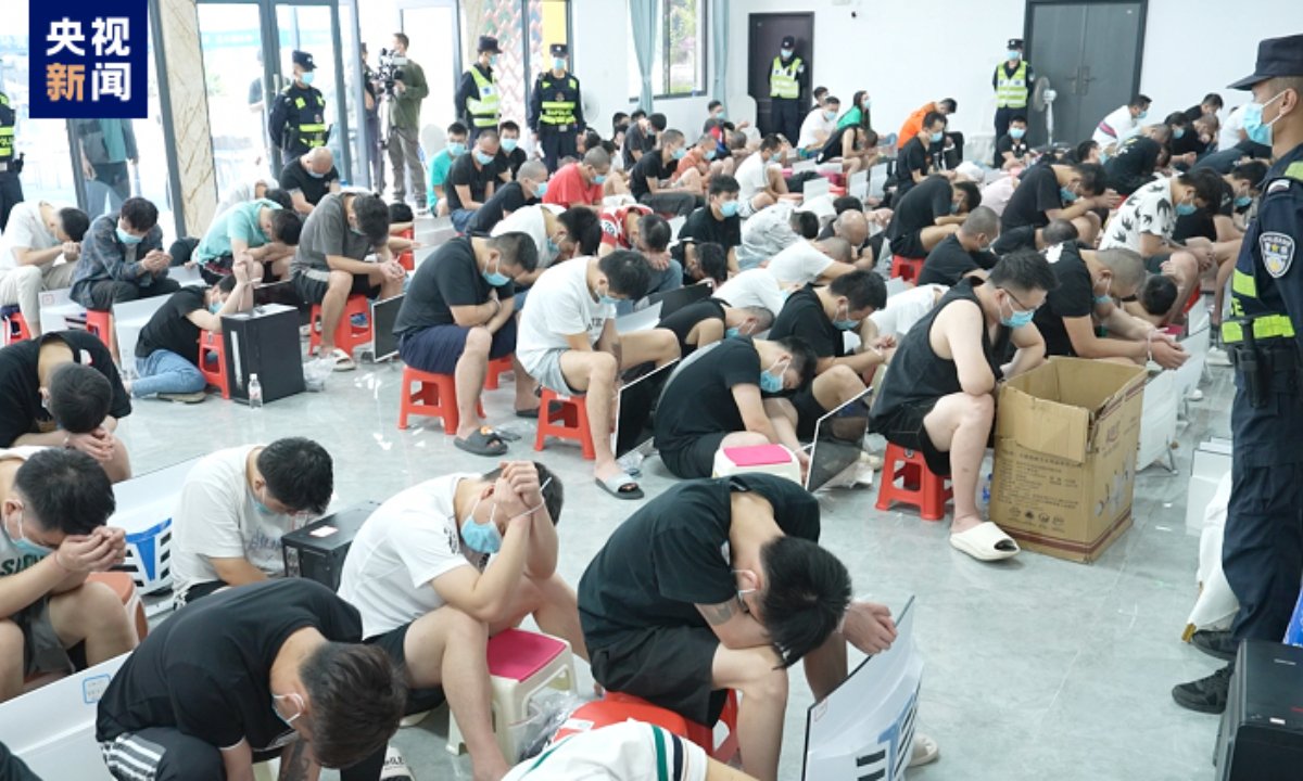 A large group of individuals seated with heads bowed, wearing masks, in a room monitored by police officers, highlighting a scene of enforcement or intervention.