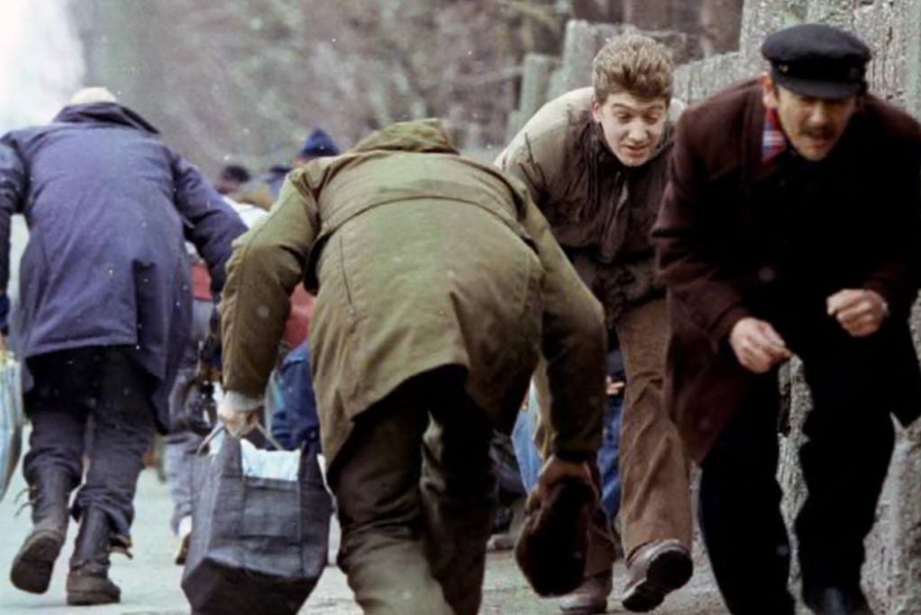 A group of people hurriedly rush down a snowy street, some carrying bags, conveying a sense of urgency and movement.