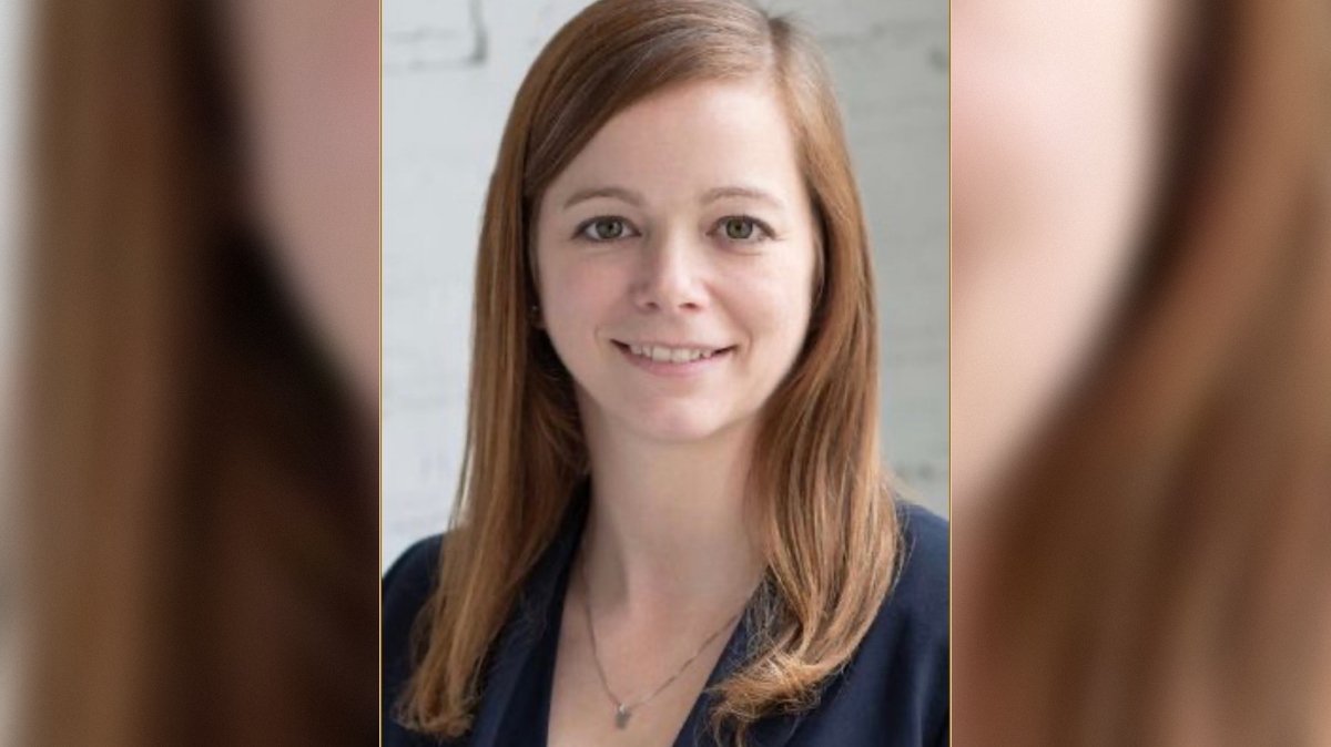 Professional headshot of a woman with shoulder-length brown hair, smiling, wearing a navy blazer against a light background.