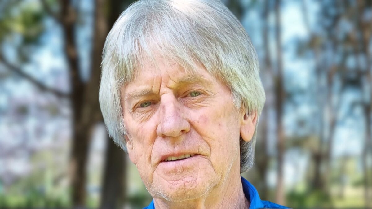 Close-up portrait of an older man with gray hair and a blue shirt, set against a blurred natural background.