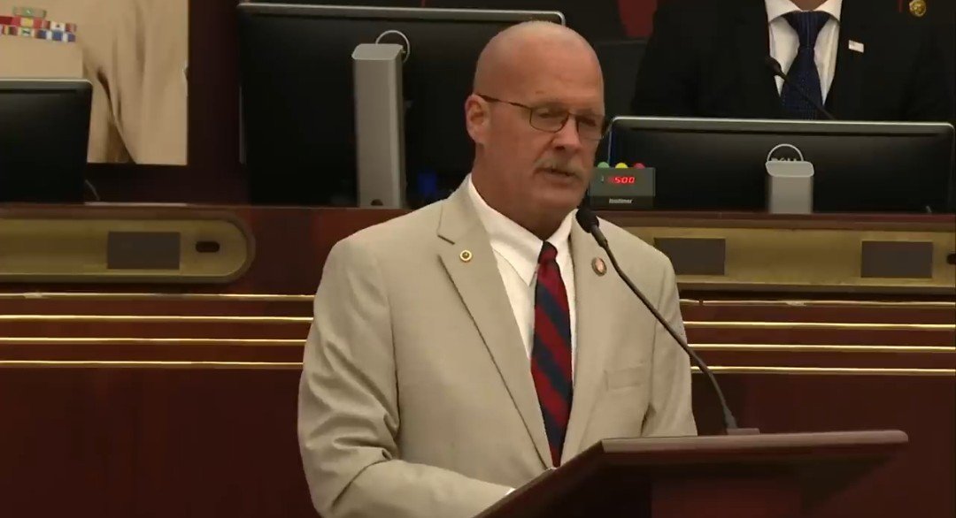 A man in a suit speaks at a podium during a formal event, with microphones and digital displays in the background.
