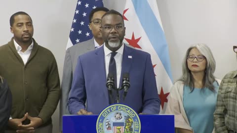 Chicago city officials gather for a press conference, featuring a speaker at the podium with the city seal, surrounded by flags and community members.