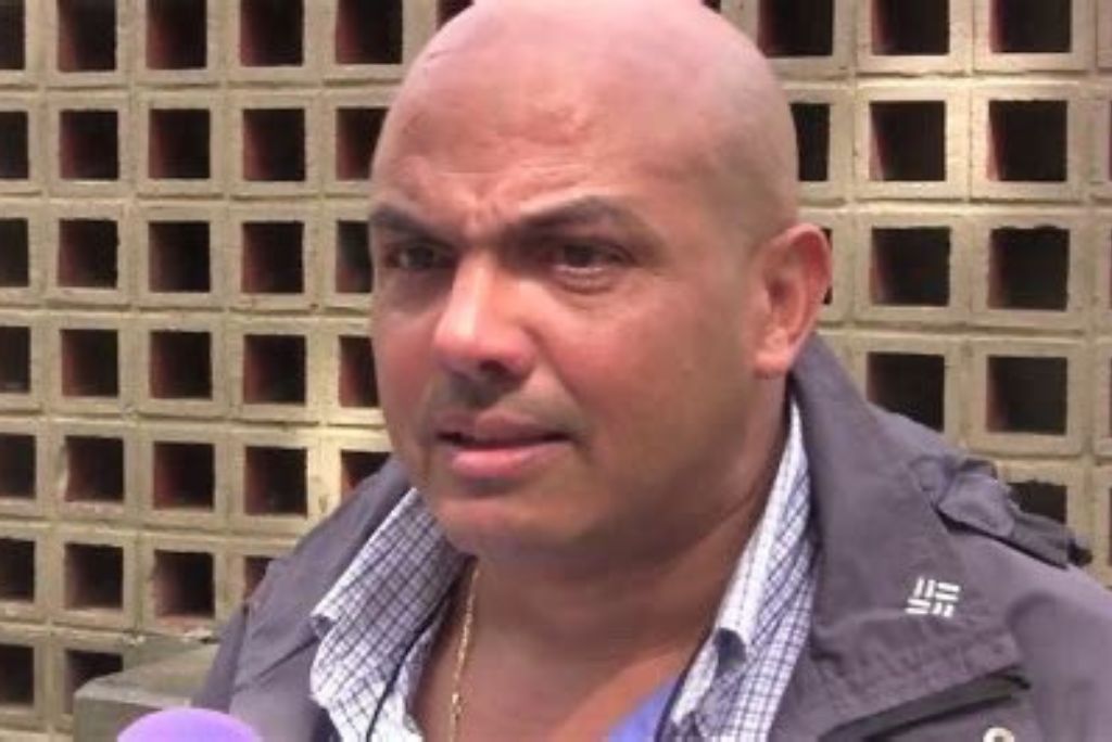 Man with a shaved head and serious expression speaking in front of a textured brick wall, wearing a jacket and plaid shirt.