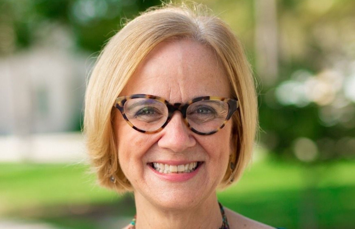 Smiling woman with short blonde hair and stylish glasses, standing outdoors against a blurred green background.