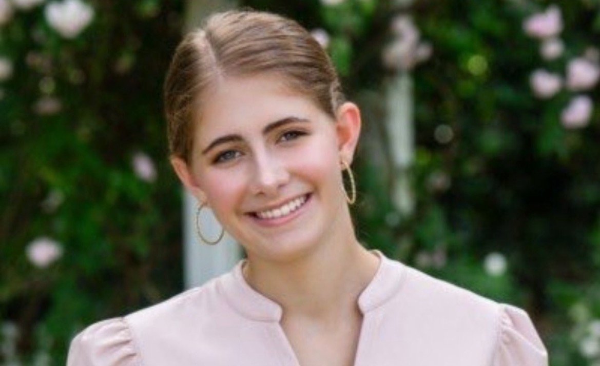 Smiling young woman with light brown hair and hoop earrings, wearing a pale pink blouse, standing in a garden with blurred flowers in the background.