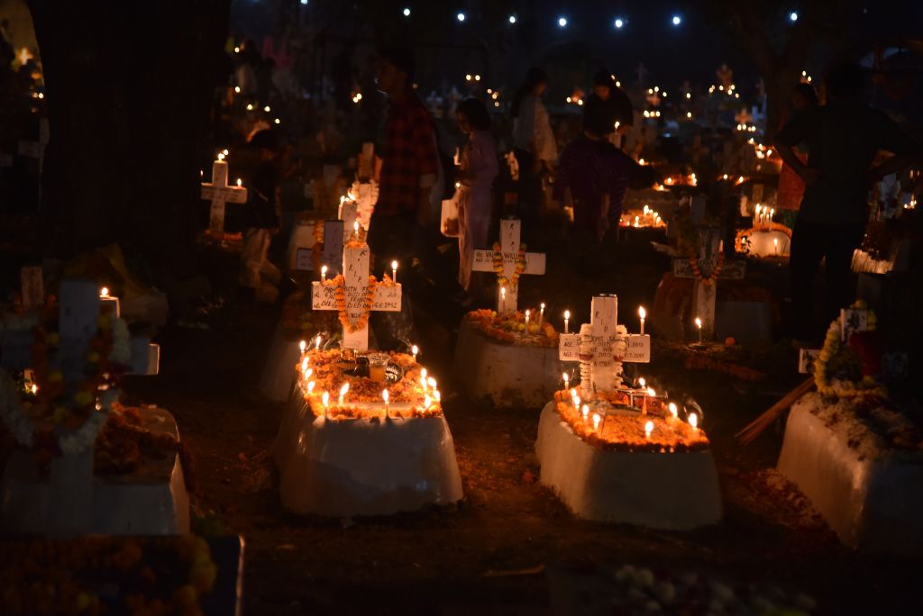 A nighttime cemetery scene illuminated by candles and flowers, with people paying their respects at graves marked by crosses.