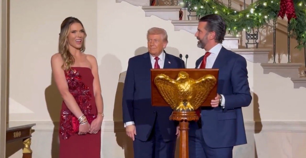 Donald Trump stands with his son and a woman in a red dress at a festive event, with holiday decorations in the background.