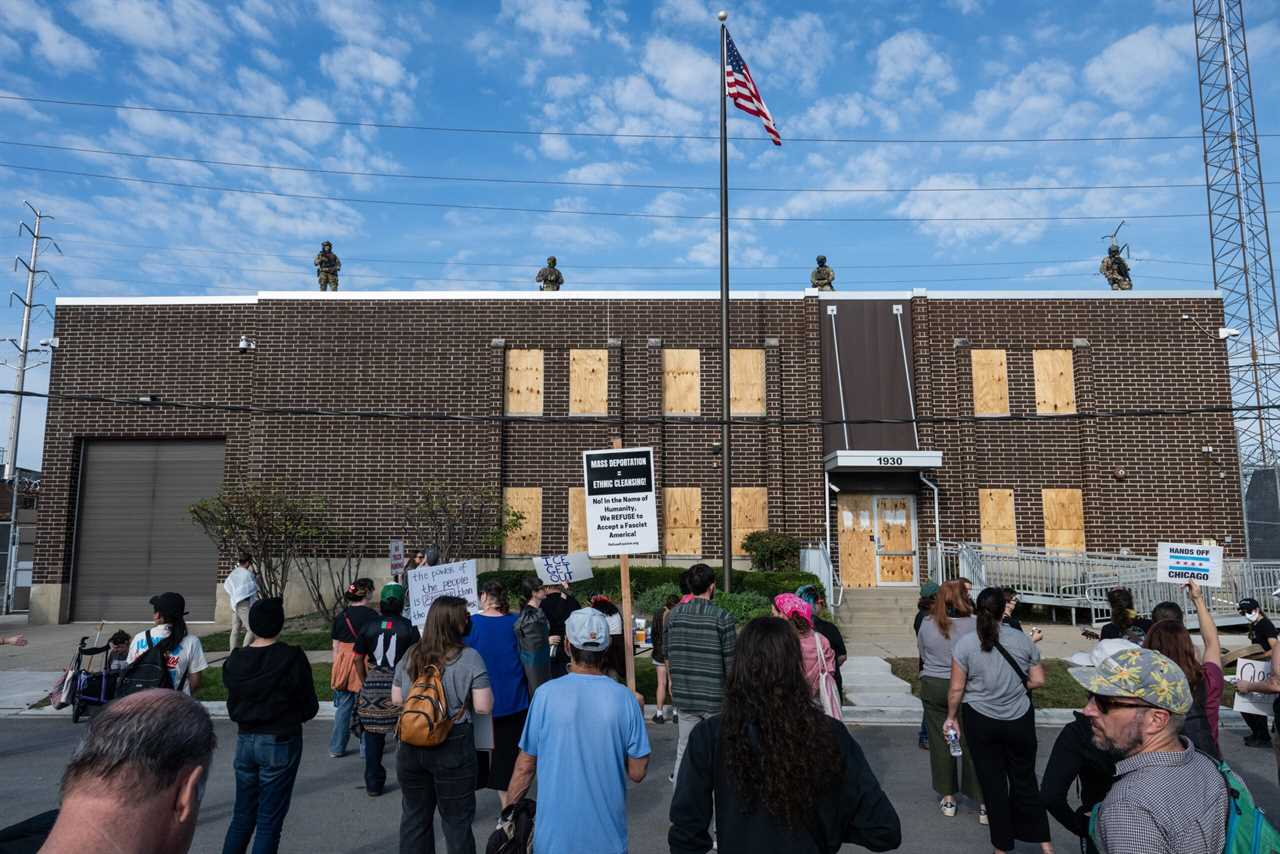 Protesters gather outside a boarded-up building with armed guards on the roof, advocating against mass deportation and expressing concerns over immigration policies.