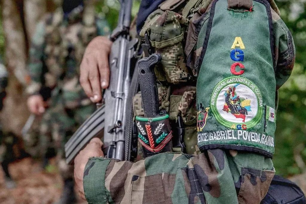 Armed soldier in camouflage uniform showcasing a patch representing the Autodefensas Gaitanistas de Colombia, with a rifle slung over his shoulder in a wooded area.