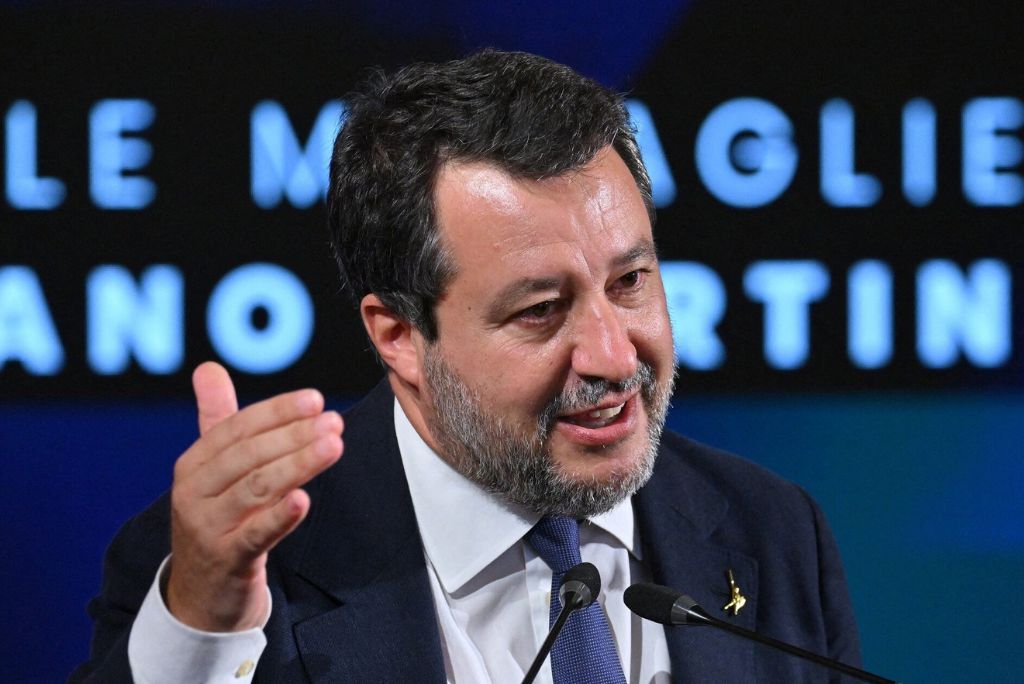 Matteo Salvini speaking at a conference, gesturing with his hand, wearing a suit, against a backdrop of illuminated text.