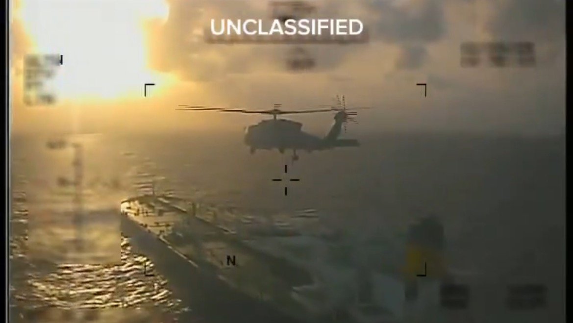 Helicopter hovering above a naval ship at sunset, captured from an aerial perspective, with an unclassified label visible.