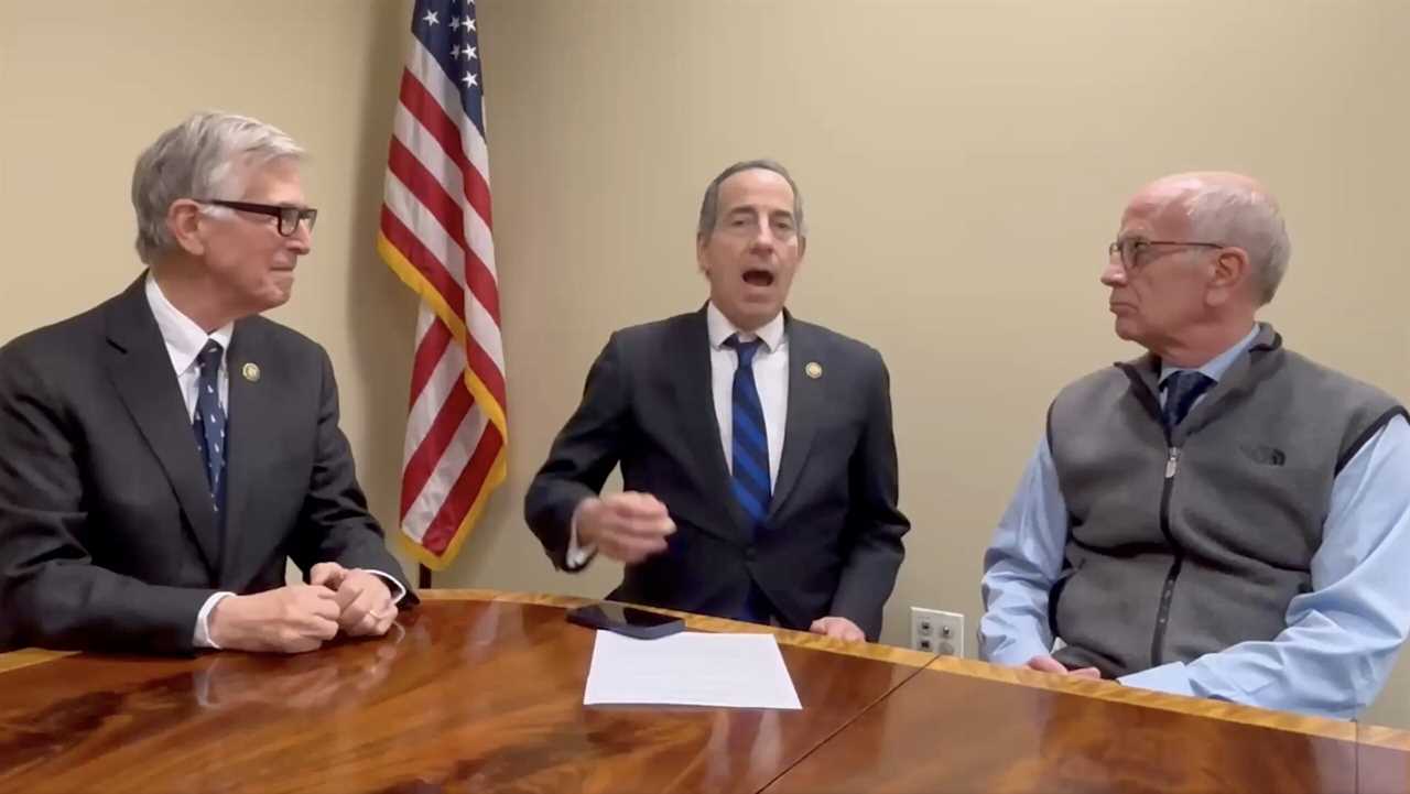 Three men in suits discussing a topic at a wooden table, with an American flag in the background.