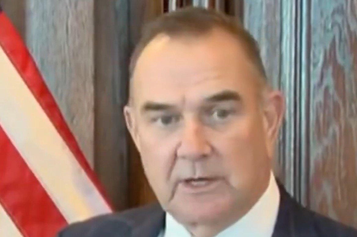 Man speaking in front of a wooden backdrop with an American flag, discussing a topic of importance in a formal setting.