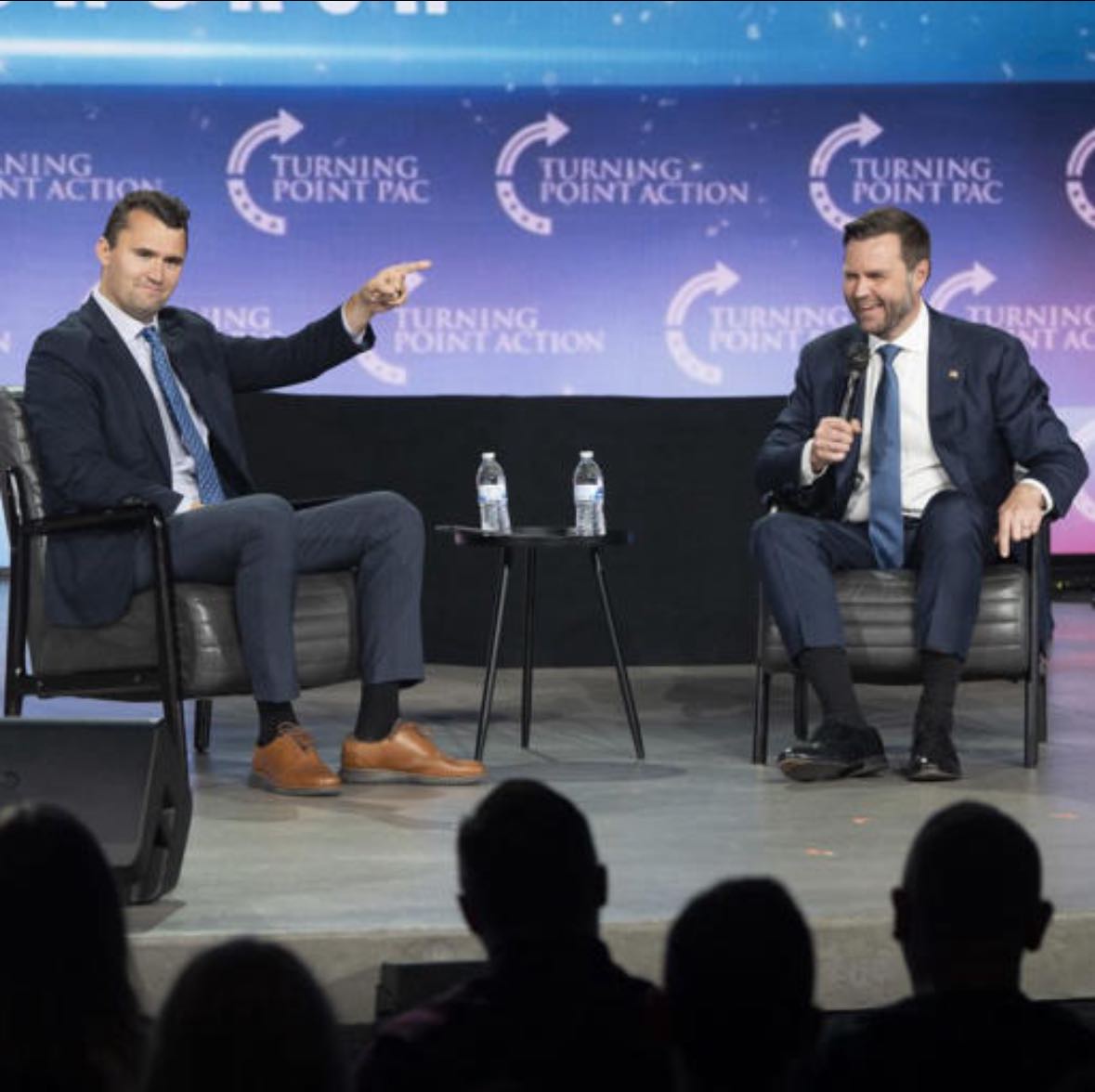 Two speakers engage in a discussion on stage at a Turning Point Action event, with an audience visible in the foreground.