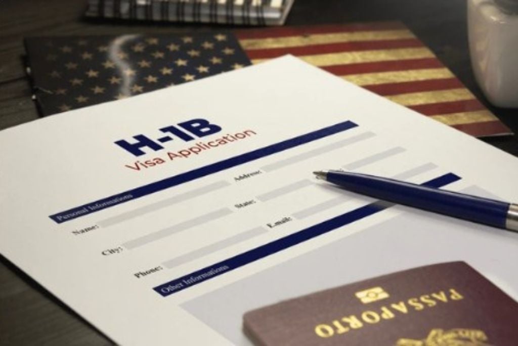 H-1B visa application form with personal information fields, a blue pen, and a passport on a wooden table with an American flag in the background.