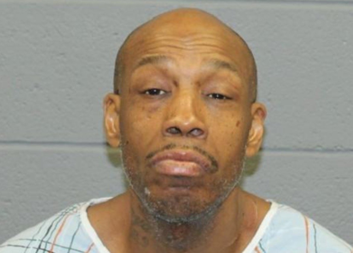 Mugshot of a man in a hospital gown with a serious expression, against a gray wall background.