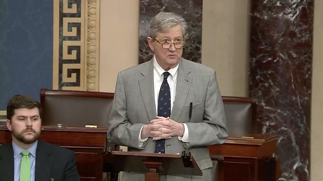A senator speaks at a podium in the Senate chamber, addressing an audience while another individual listens attentively in the foreground.