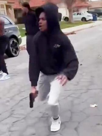 Individual in a hoodie crouching on a street holding a firearm, with other figures in the background, capturing a tense urban scene.