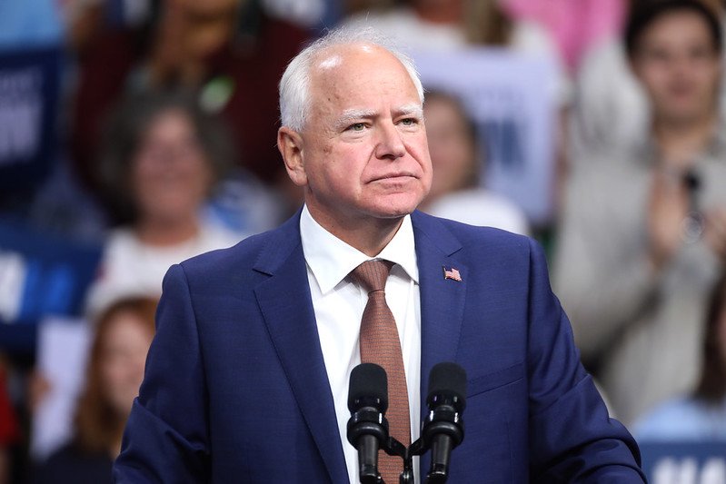 Minnesota Governor Tim Walz speaks at a rally, addressing an engaged crowd with campaign signs in the background.