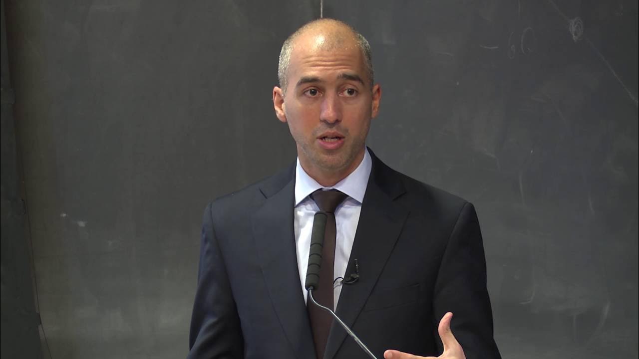 Business professional giving a presentation, wearing a suit and tie, with a dark background and microphone in front.