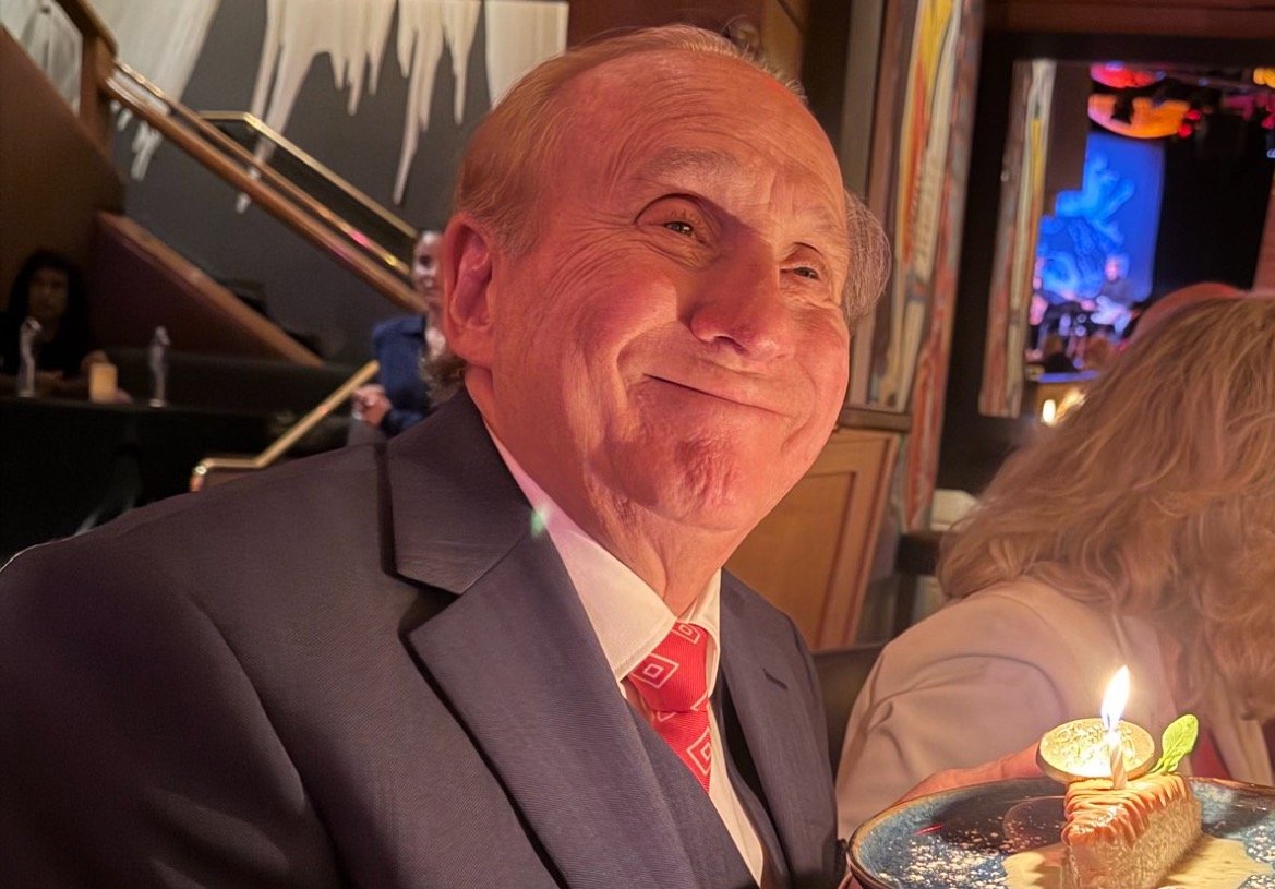 Smiling man in a suit holding a dessert with a candle at a festive gathering, surrounded by a lively atmosphere.