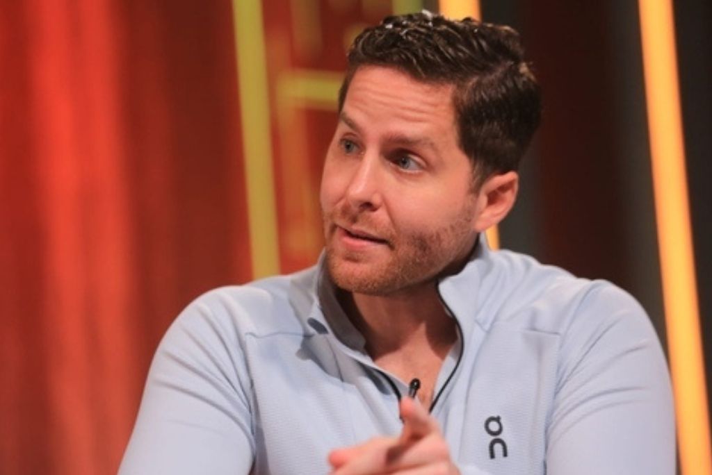 Man in a light blue athletic shirt engaged in conversation, with a blurred red background, showcasing an expressive gesture while speaking.