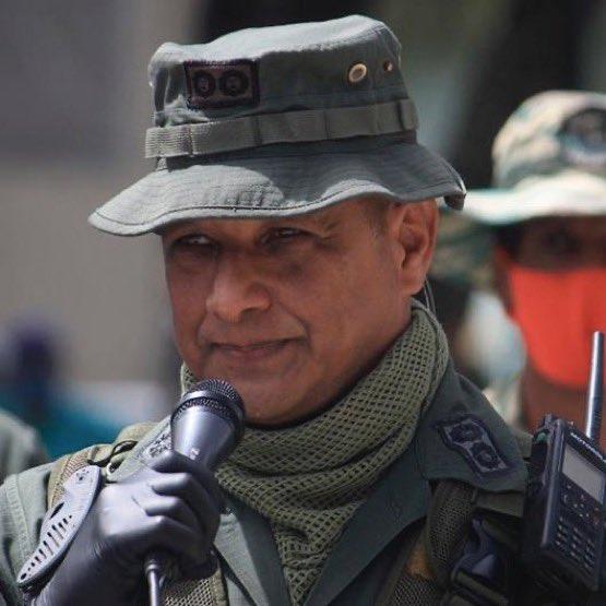 Military officer speaking into a microphone while wearing a hat and tactical gear during a public event.