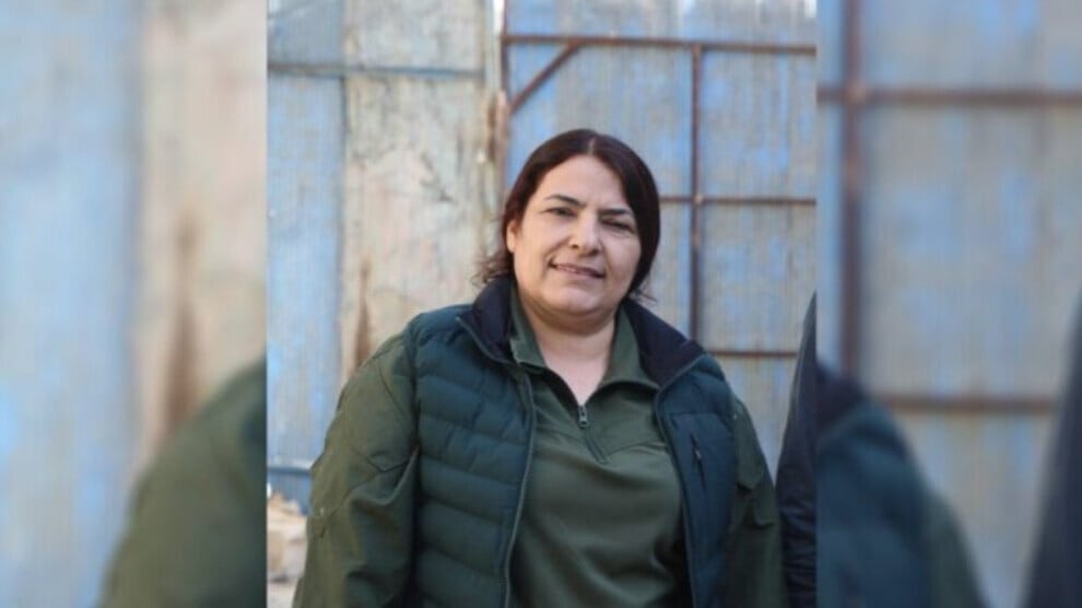 Woman wearing a green jacket smiling in front of a blue and brown industrial background.